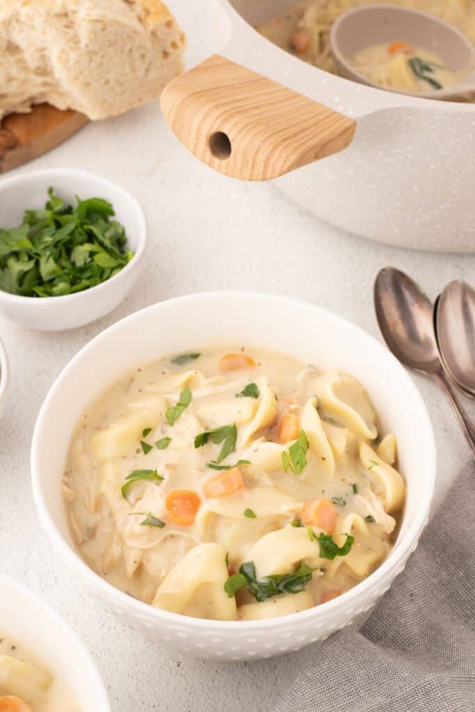 White bowl of soup. Large pot of soup in the background and with a loaf of bread, cutting board and small bowl of parsley.