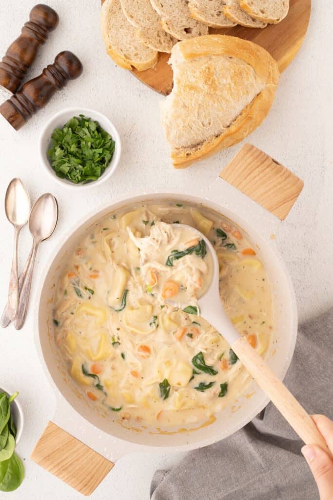 Large pot of chicken tortellini soup with a ladle lifting a serving up. Pot is surrounded by spinach leaves, parsley, salt and pepper shakers, and sliced bread on a cutting board.