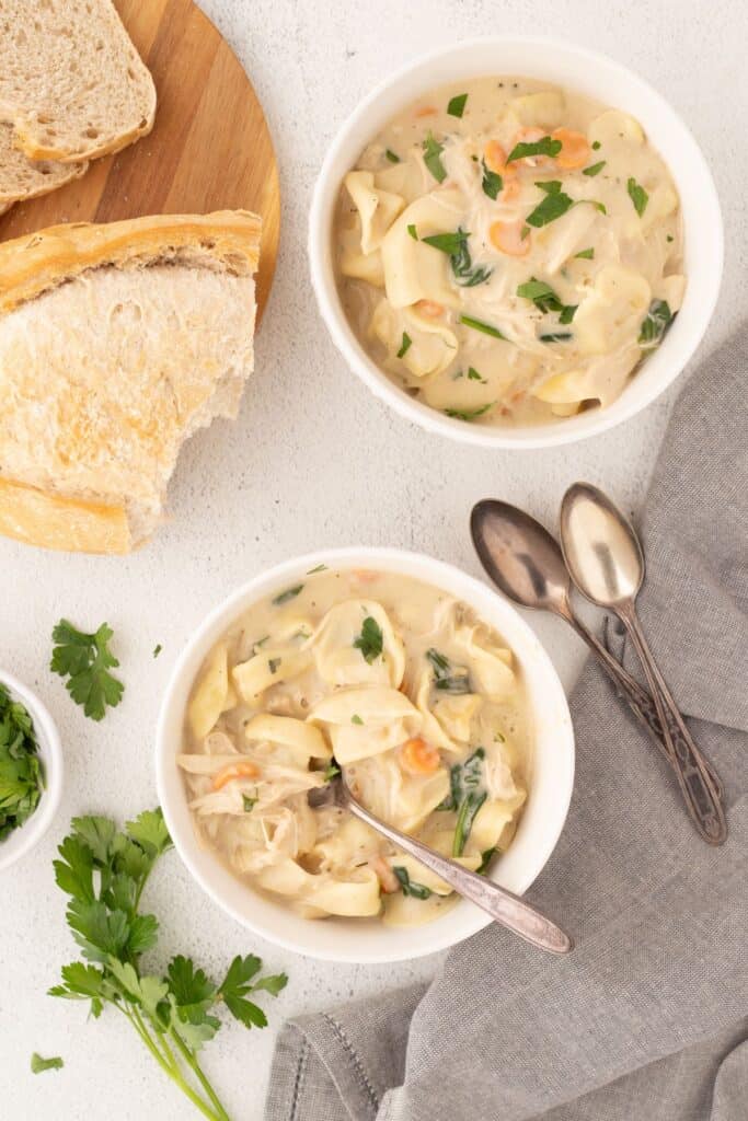 Two white bowls of chicken tortellini soup with spoons. To the left is a loaf of sliced bread on a cutting board.