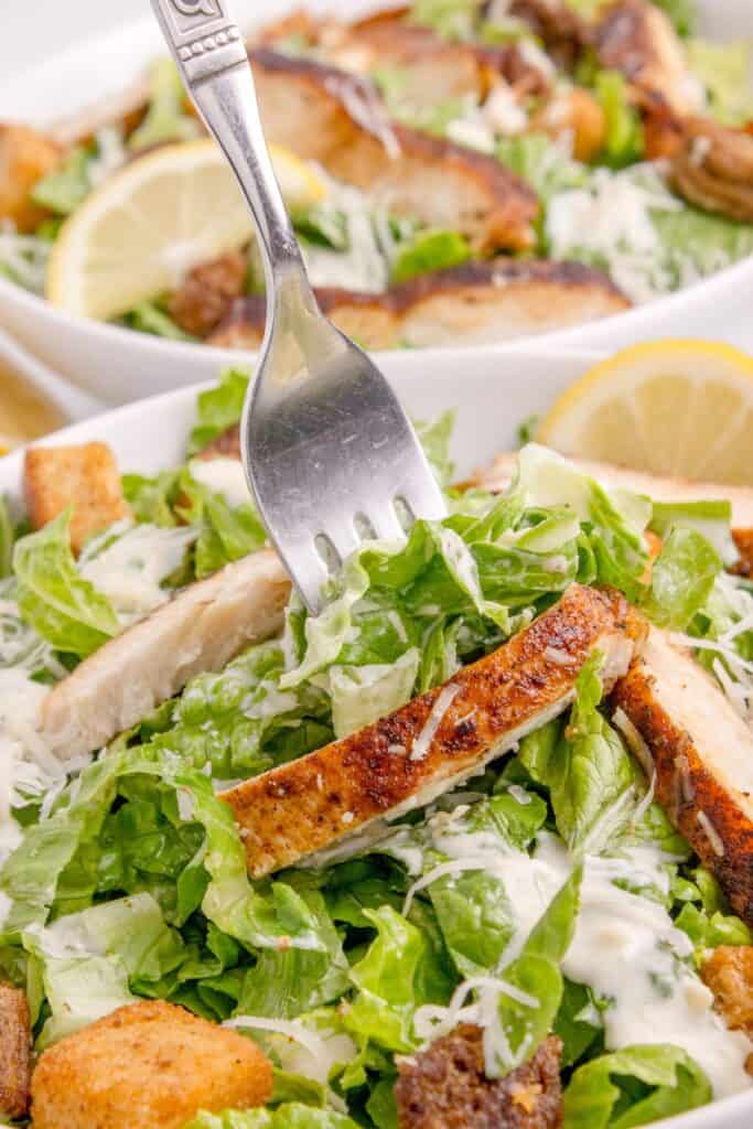 Two dressed chicken caesar salads. The salad in the foreground has a fork lifting a bite from the bowl.