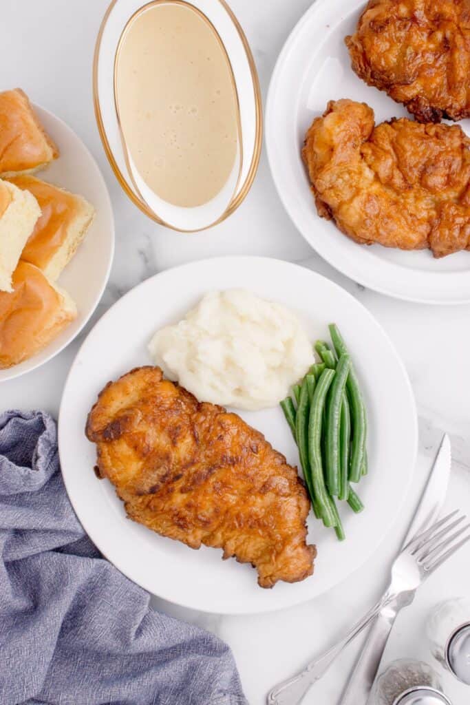 Above view of fried chicken on a dinner plate with mashed potatoes, green beans, and fried chicken. Plate is surrounded by additional plates of fried chicken, a gravy boat with gravy, and dinner rolls.