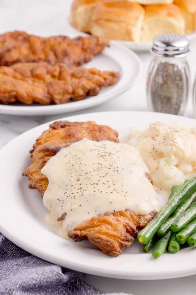 White dinner plate with green beans, mashed potatoes, and chicken fried chicken with gravy. Another plate of fried chicken is in the background with a plate full of dinner rolls.