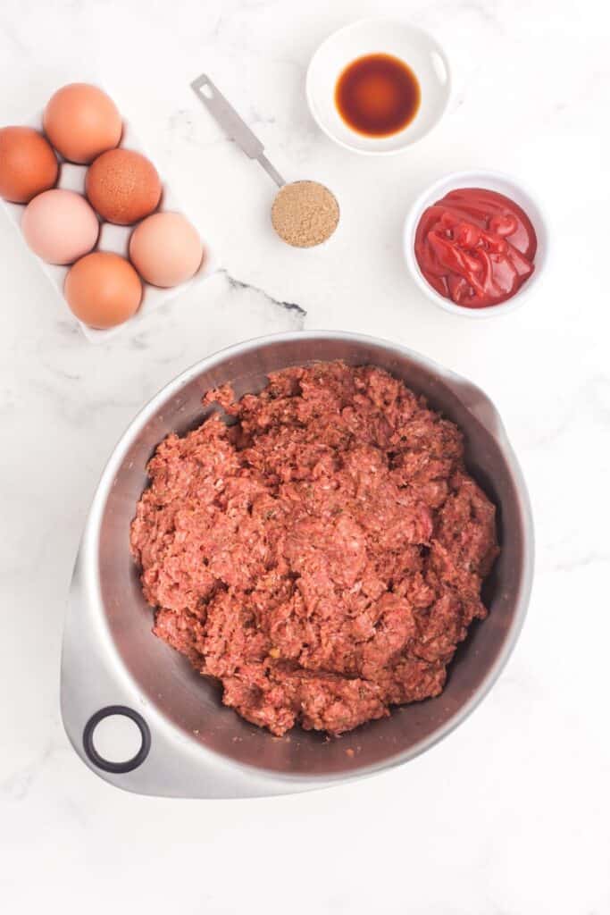 Ground beef in a metal mixing bowl. Bowl is surrounded by uncracked eggs, spoon full of brown sugar, and small bowls of worcestershire sauce, and ketchup.