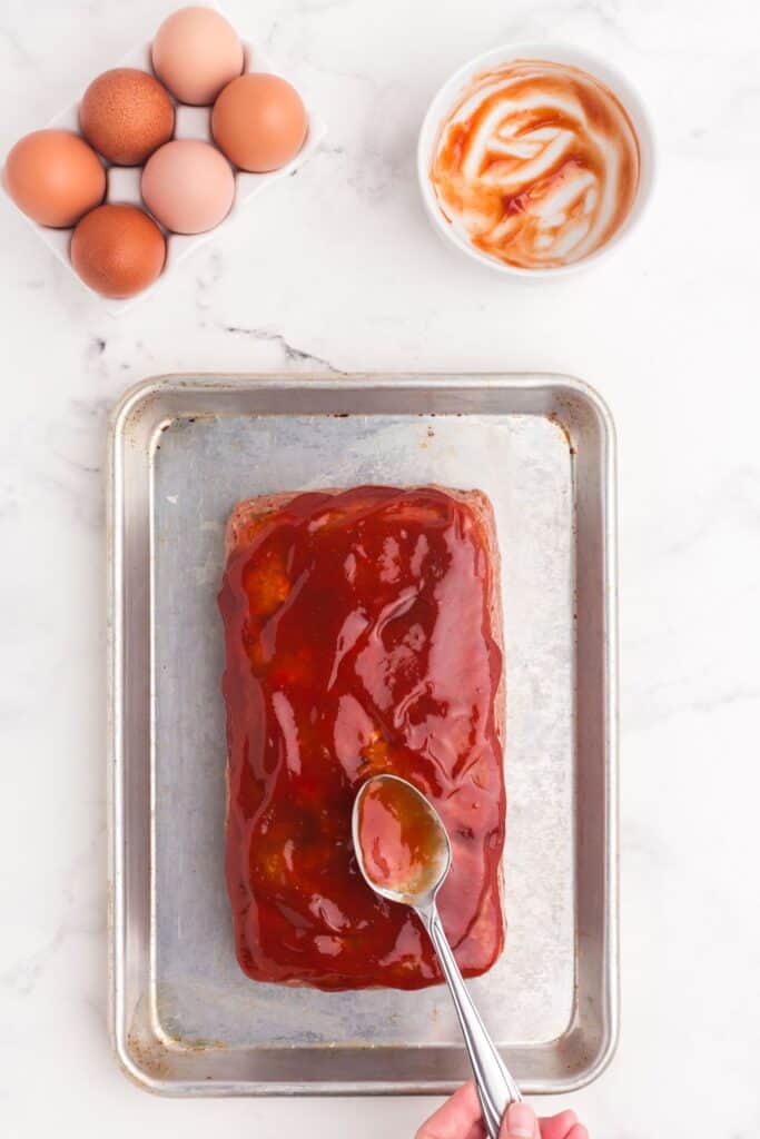 Uncooked meatloaf on a baking sheet, with a spoon smoothing over the ketchup topping.