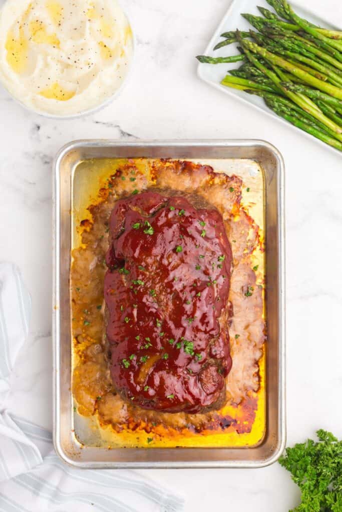 Baking sheet with baked meatloaf on it. Bowl of mashed potatoes shown at the top left, and a plate a asparagus spheres on the top right.