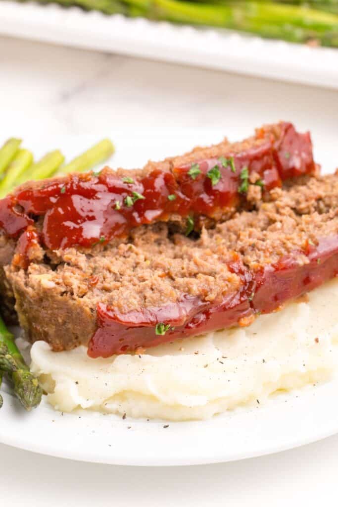 Close up of white dinner plate with asparagus, mashed potatoes, and meatloaf layered on top of the potatoes.