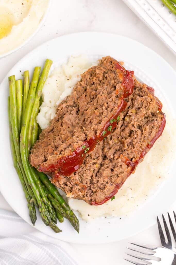 White dinner plate with asparagus, mashed potatoes, and meatloaf layered on top of the potatoes.