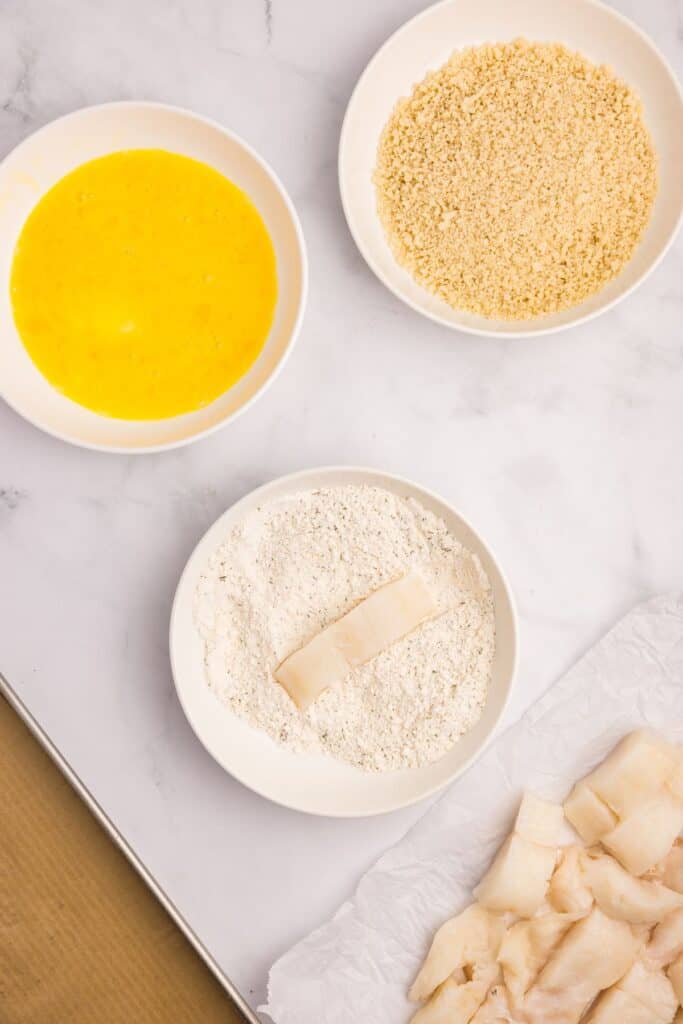 Three bowls prepped as a breading station. Top bowl has bread crumbs, middle bowl has a whisked egg, bottom bowl has seasoned flour. Below the bowls are unbreaded fish pieces.