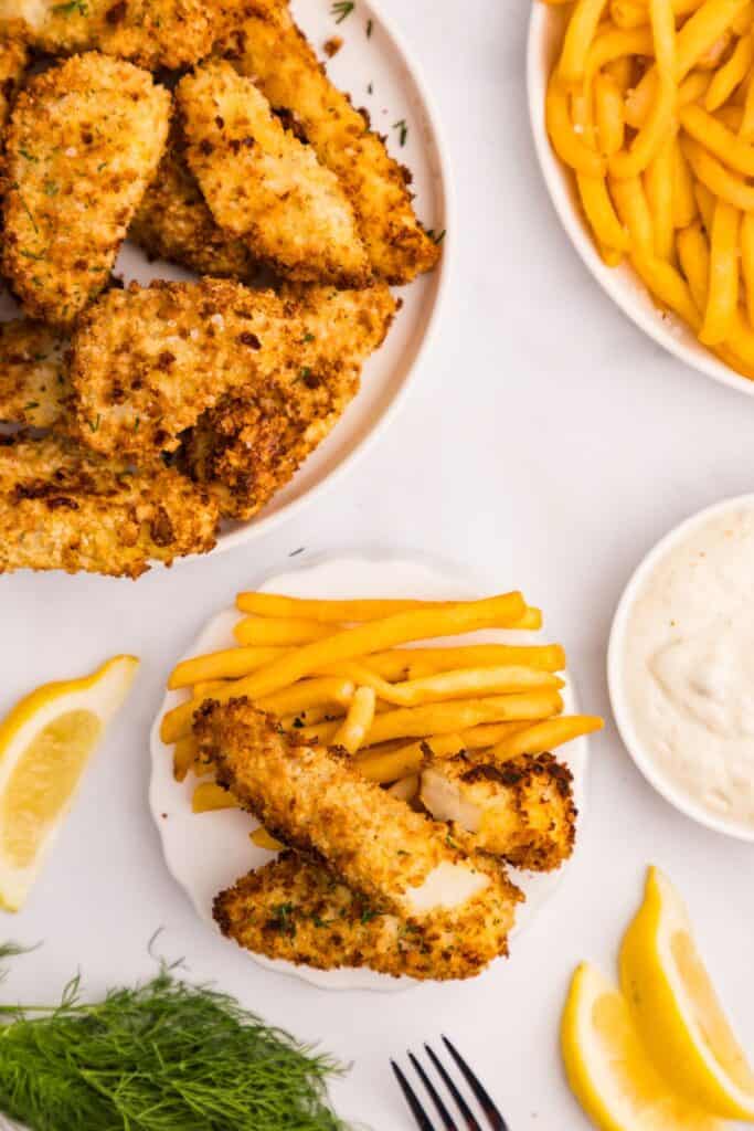 White white dinner plate piled high with fish sticks and french fries. Surrounded by a large platter of fish sticks, a platter of french fries, a bowl of tartar sauce, lemons and fresh dill.