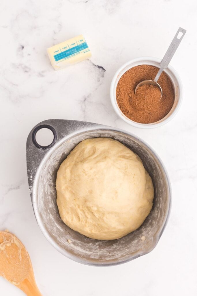 Metal mixing bowl with cinnamon roll dough next to a small bowl with cinnamon and sugar mix, and a half stick of butter.