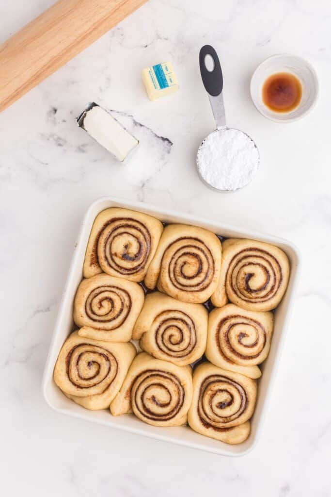 Baked cinnamon rolls in a baking pan surrounded by ingredients for icing.