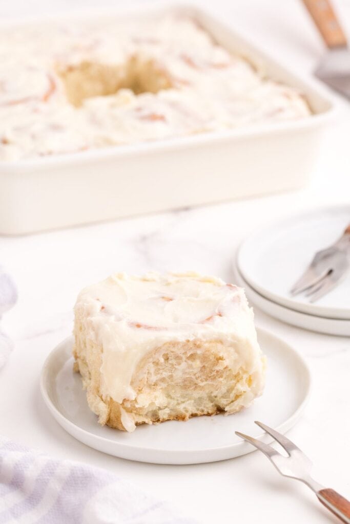 Baked and iced cinnamon roll on a white plate. The baking dish with the rest of the cinnamon rolls is in the background.