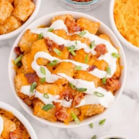 Loaded tater tots in a white bowl surrounded by the ingredients in small bowls: Tater tots, crumbled bacon, cheddar cheese, and green onions.