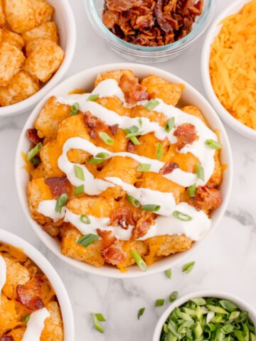 Loaded tater tots in a white bowl surrounded by the ingredients in small bowls: Tater tots, crumbled bacon, cheddar cheese, and green onions.