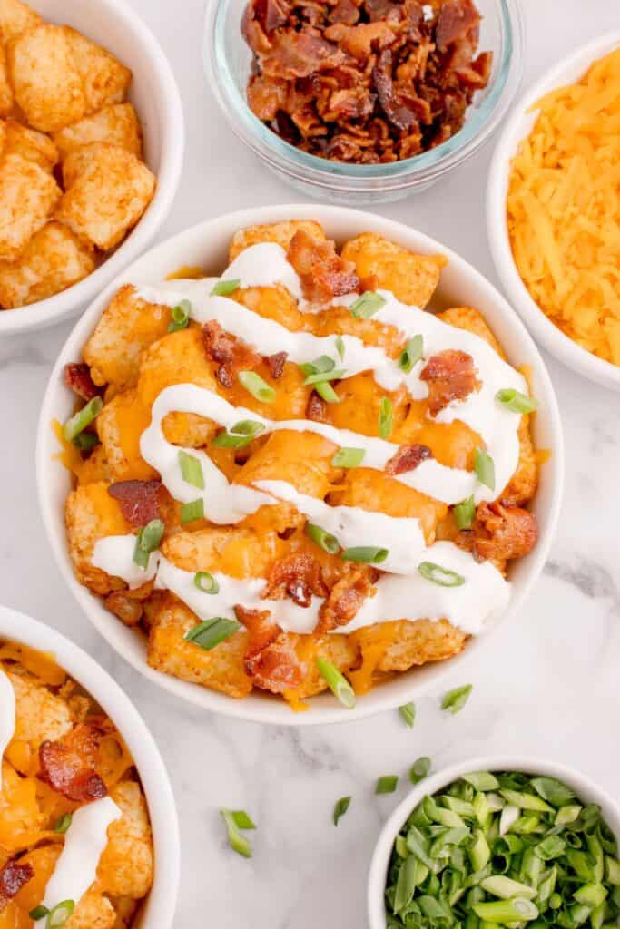 Loaded tater tots in a white bowl surrounded by the ingredients in small bowls: Tater tots, crumbled bacon, cheddar cheese, and green onions.