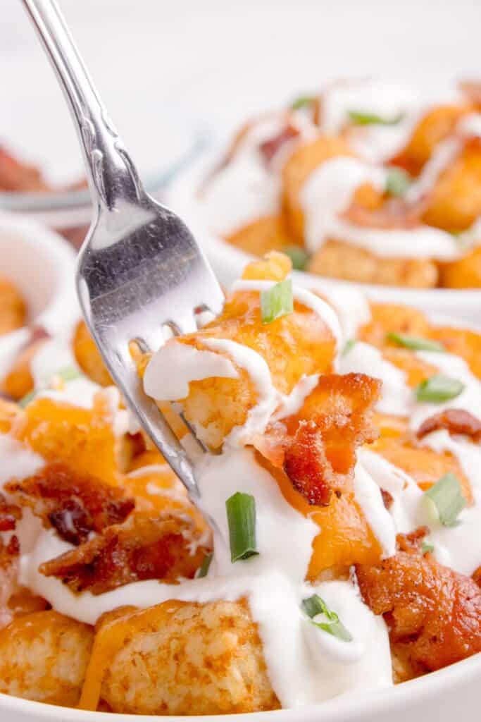 Close up of tater tots with sour cream, bacon, cheese, and green onions. A fork is lifting a bite.