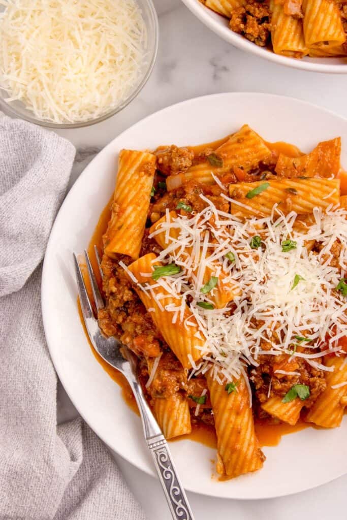 A white dinner plate with pasta bolognese and a fork. Above the plate is small glass bowl of grated parmesan cheese.