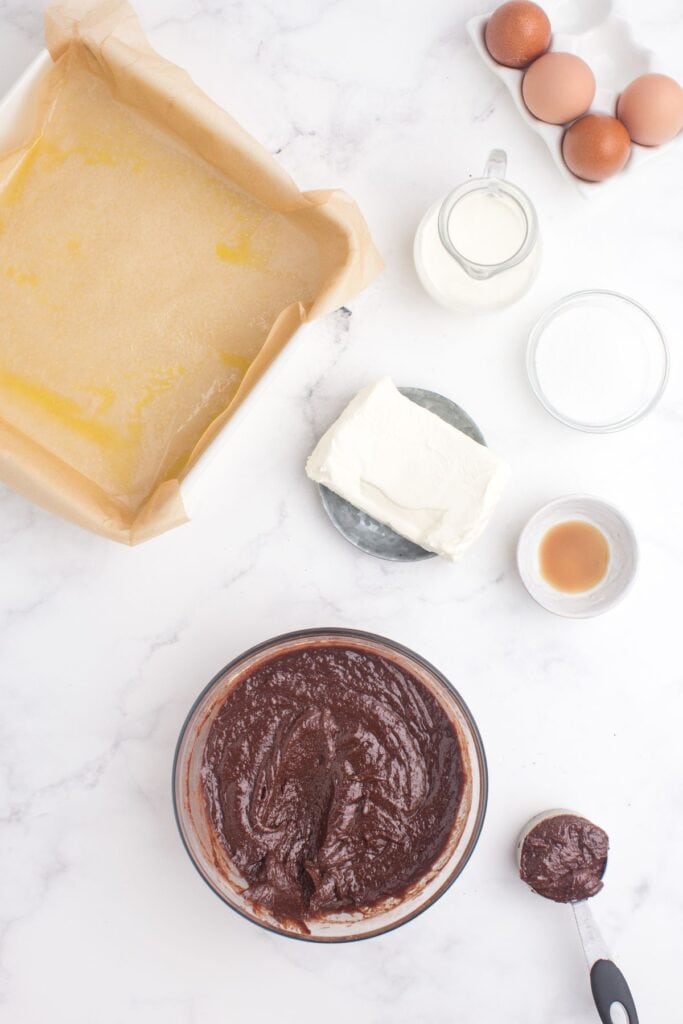 Brownie batter is prepped in a glass bowl. One small measuring cup has a some brownie batter in it. Baking pan is prepped with parchment. Ingredients for the cream cheese layer are prepped in the picture.