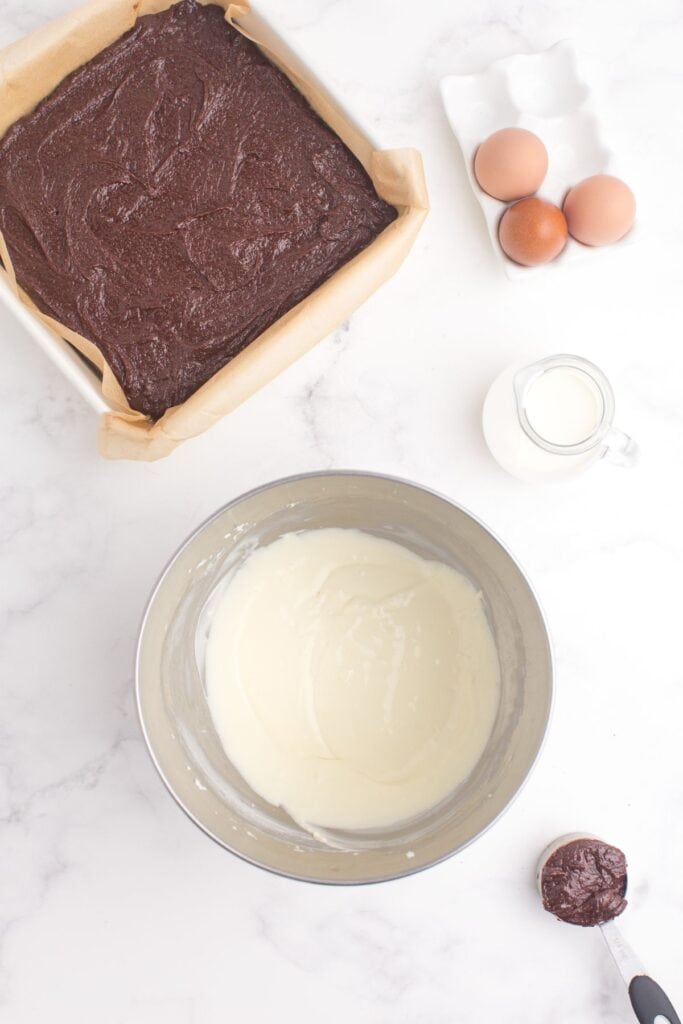 Baking pan with brownie batter in it next to three whole eggs and a pitcher of milk. A separate bowl with the cream cheese mixture is prepped with a small measuring cup full of brownie batter.