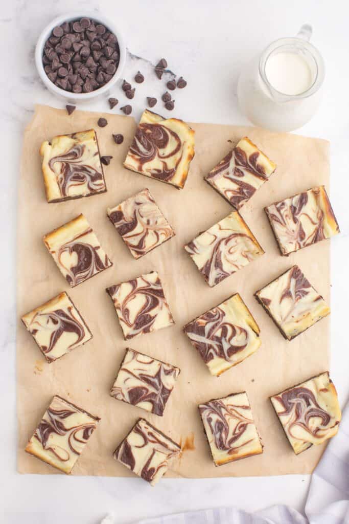 Cheesecake brownies cut and spread out on a sheet of parchment paper. A small bowl of chocolate chips and a pitcher of milk are placed above the parchment.