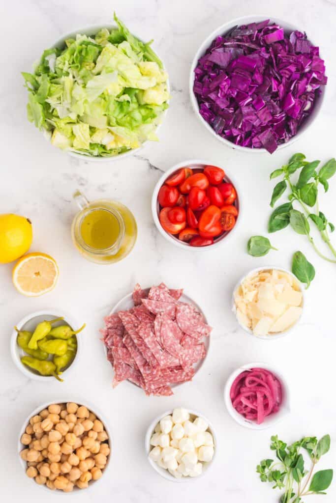 Ingredients for chopped salad: iceberg lettuce, red cabbage, pickled red onion, cherry tomatoes, chickpeas, mozzarella pearls, provolone, pepperoncini, oregano, basil, salami, extra virgin olive oil, fresh lemon juice, red wine vinegar, Dijon mustard, dried oregano, garlic powder, onion powder, salt, and pepper.