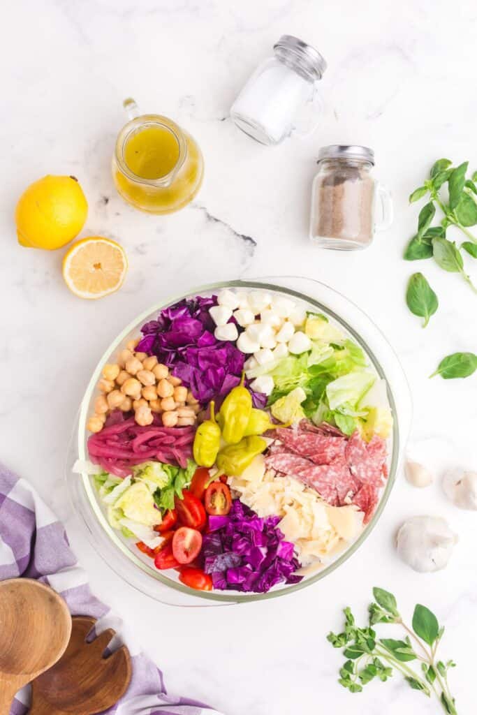 Chopped salad in a glass bowl surrounded by a pitcher of salad dressing, lemons, fresh basil, fresh oregano, garlic, salad tongs, salt, and pepper.