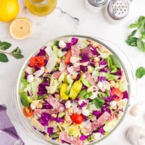 Chopped salad in a glass bowl surrounded by a pitcher of salad dressing, lemons, salad tongs, salt, and pepper.