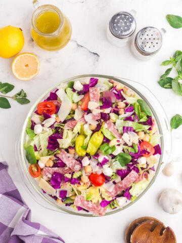 Chopped salad in a glass bowl surrounded by a pitcher of salad dressing, lemons, salad tongs, salt, and pepper.