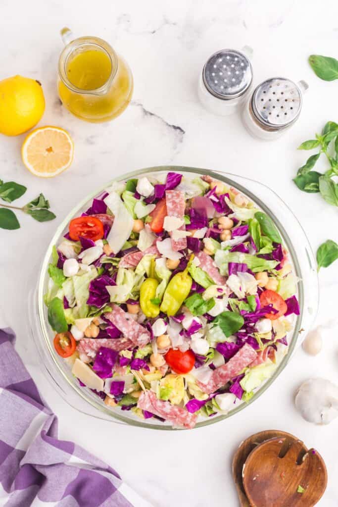 Chopped salad in a glass bowl surrounded by a pitcher of salad dressing, lemons, salad tongs, salt, and pepper.