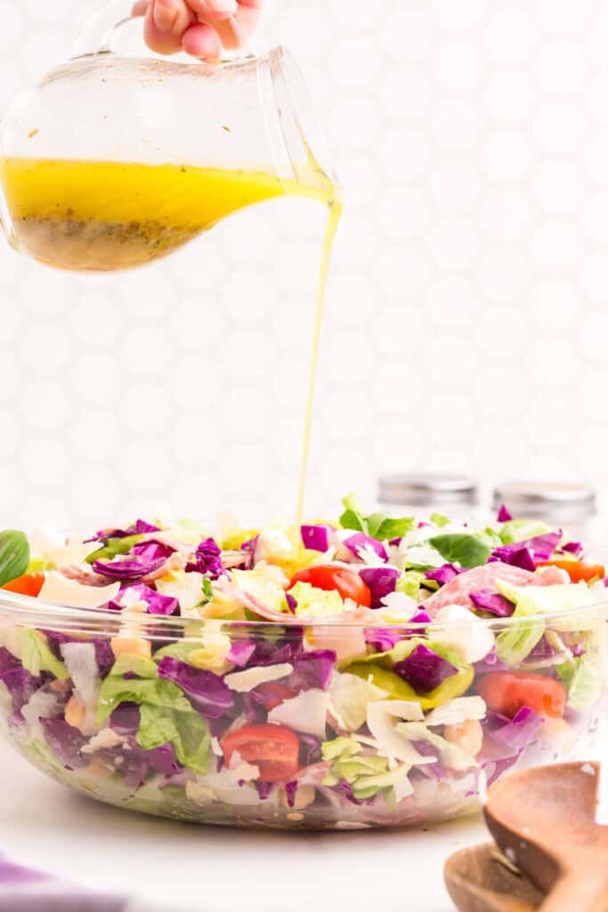 Glass bowl with chopped salad. A pitcher is pouring the dressing on the salad.