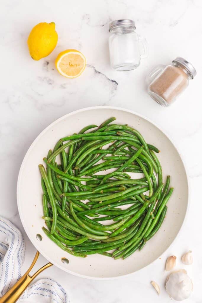 Cooked garlic green beans in a skillet surrounded by a fresh head of garlic, lemon, salt and pepper shakers.