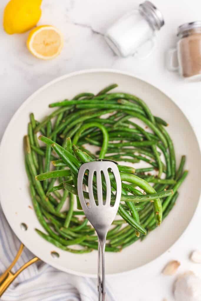 Completed garlic green bean recipe. A pair of serving tongs are lifting a serving above the pan.