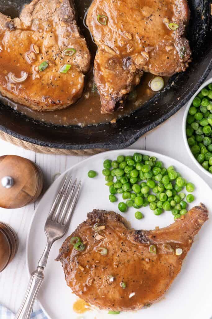 White dinner plate with a pork chop and serving of peas. Next to the plate is a cast iron pan with pork chops in it.