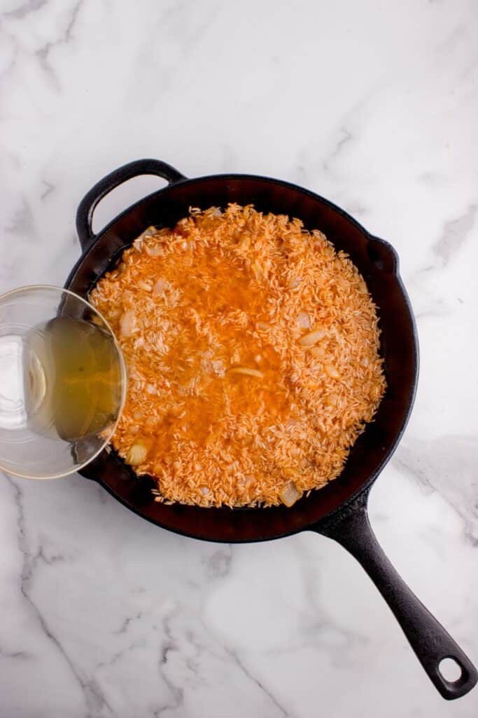 Rice in a cast iron pan. A glass bowl of chicken broth is poured into the pan.