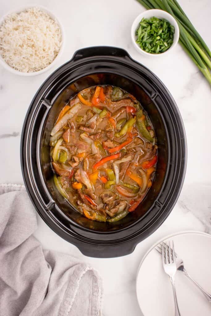 Completed slow cooker pepper steak in the crock pot surrounded by a bowl of white rice, a bowl of chopped green onions, plates, and forks.