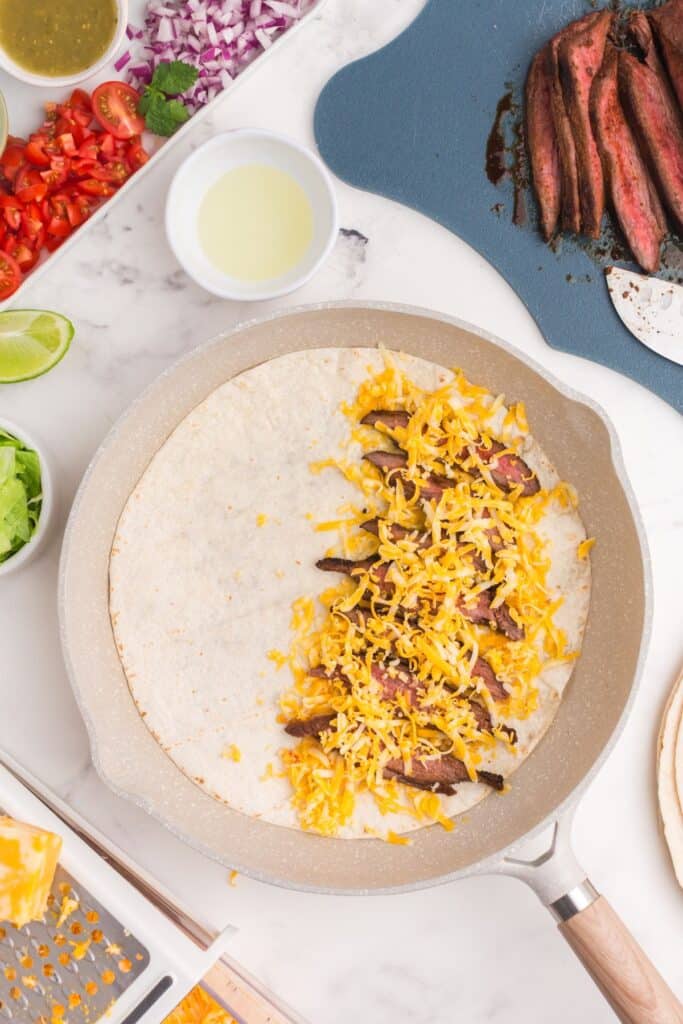 Open tortilla in a skillet loaded with sliced steak and shredded cheese.