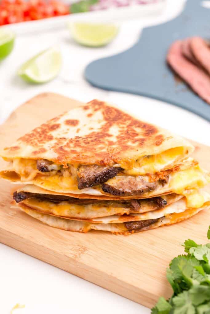 Stacked quesadillas on a cutting board surrounded by fresh cilantro, a cutting board with steak on it, sliced limes, and a tray of chopped onions and tomatoes.