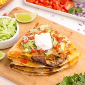 Stacked quesadillas on a cutting board. Topped with lettuce, tomatoes, and sour cream.