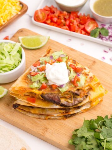 Stacked quesadillas on a cutting board. Topped with lettuce, tomatoes, and sour cream.