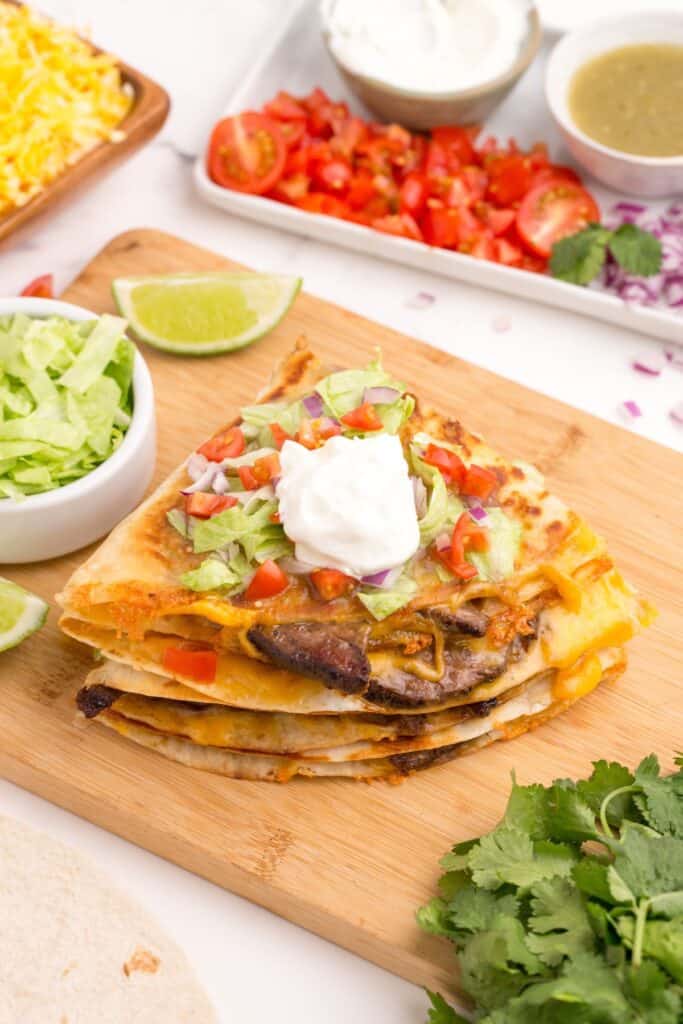 Stacked quesadillas on a cutting board. Topped with lettuce, tomatoes, and sour cream.