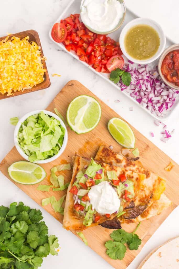 Steak quesadilla on a cutting board with chopped limes. Surrounded by fresh cilantro, salsa, hot sauce, sour cream, shredded cheese, chopped tomatoes and chopped onions.