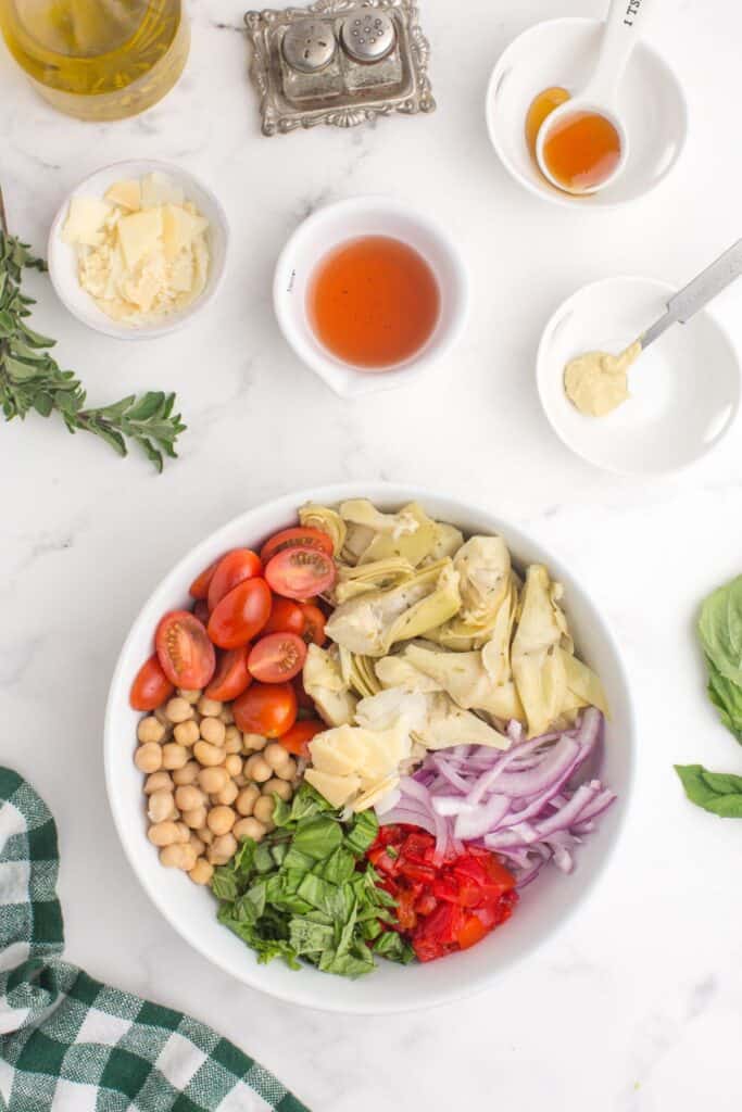 Assembled artichoke salad in a serving bowl. Above the bowl are the ingredients needed for the dressing in small bowls.