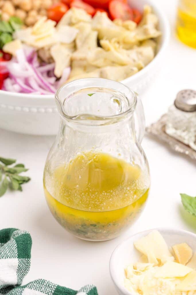 Small glass pitcher of homemade salad dressing. A serving bowl of artichoke salad is in the background.