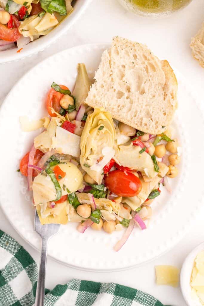 A white dinner plate with artichoke salad and a slice of artisan bread on it.