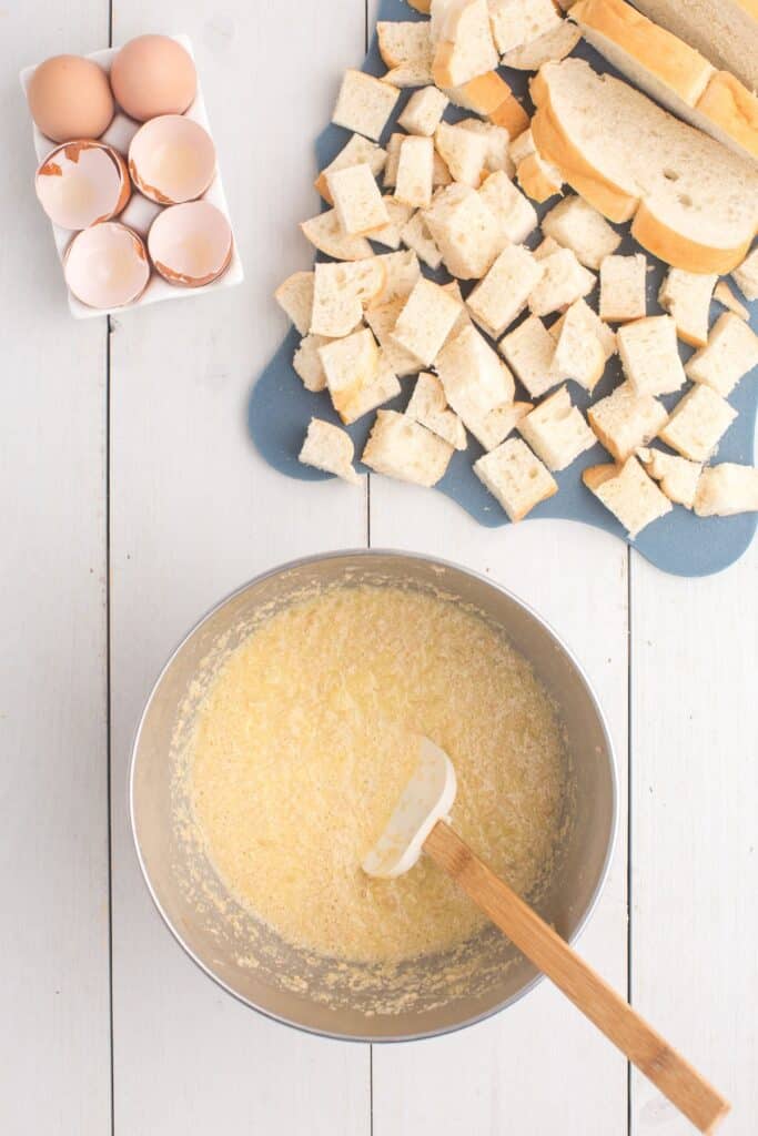 Mixing bowl with butter, sugar, salt, eggs, cinnamon, and crushed pineapple. A cutting board next to the bowl has cubed bread chunks.