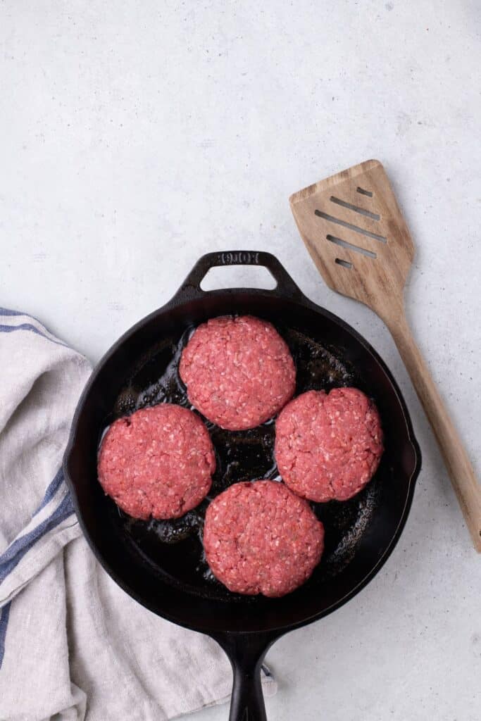 Raw ground beef patties being cooked in a cast iron pan.