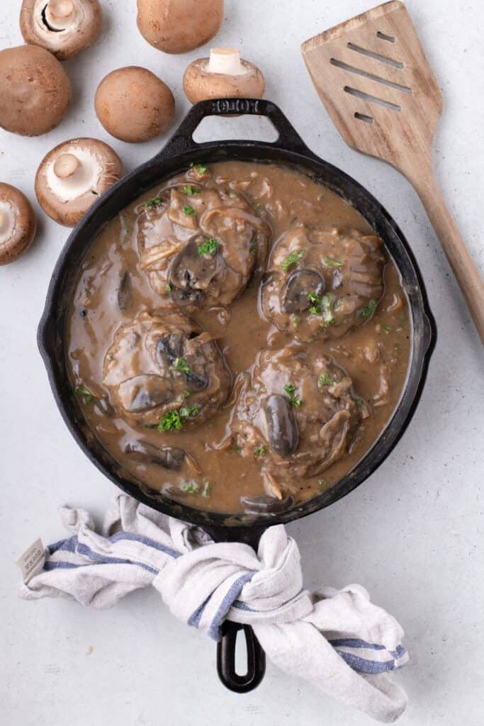 Cast iron pan with cooked hamburger steak in a gravy. Pan is surrounded by a bunch of mushrooms and a wooden spatula.