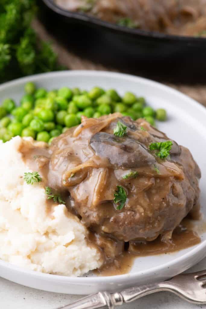 White dinner plate with hamburger steak. Served with mashed potatoes and peas.