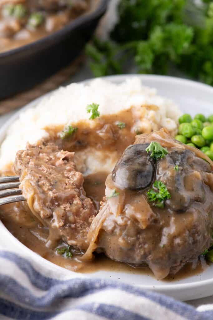 Serving of hamburger steak on a white dinner plate, served with mashed potatoes and peas.